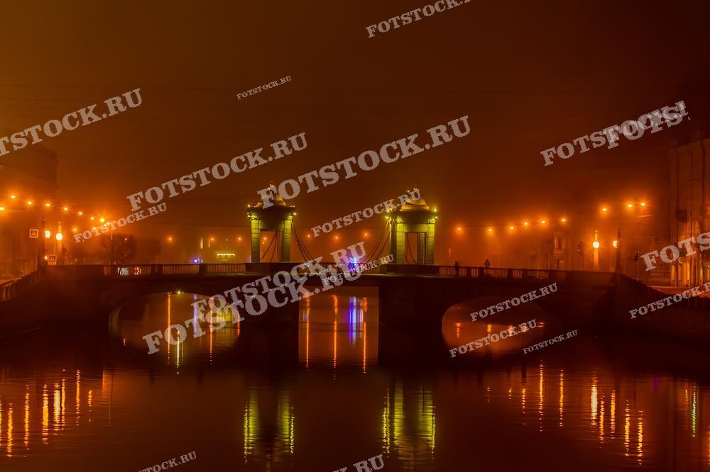 Фотография изображает мост Ломоносова в Санкт-Петербурге, окутанный ночным туманом. Фонари создают мягкое свечение, которое отражается в водах реки, придавая изображению мистический и загадочный характер. Такая атмосфера подходит для использования в материалах, акцентирующих внимание на уникальной культуре и атмосфере Петербурга, будь то художественные проекты, фотоэкспозиции или публикации о городе.